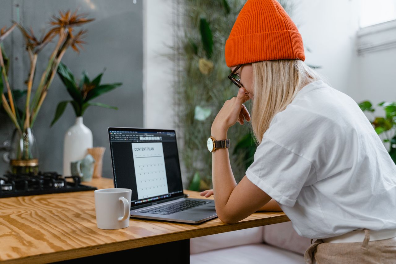 Young woman in orange beanie working on a laptop with a content plan in a cozy home office setting.